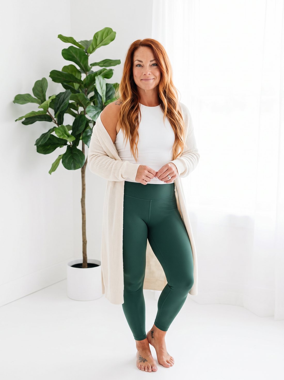 Ashley in a cream cardigan and green leggings beside a plant